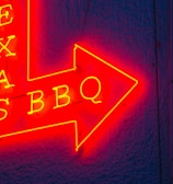A vibrant red and yellow neon sign in the shape of an arrow pointing to the right, featuring the words 'TEXAS BBQ' with a textured dark blue wall background.