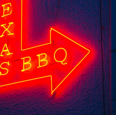 A vibrant red and yellow neon sign in the shape of an arrow pointing to the right, featuring the words 'TEXAS BBQ' with a textured dark blue wall background.