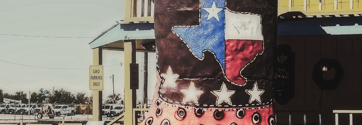A large decorative cowboy boot with the text 'Big Texan' is displayed outdoors. The boot features a design with eye-catching colors and patterns, including a depiction of the Texas flag and stars. The setting includes a bright yellow building with western-style architectural elements in the background.