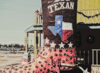 A large decorative cowboy boot with the text 'Big Texan' is displayed outdoors. The boot features a design with eye-catching colors and patterns, including a depiction of the Texas flag and stars. The setting includes a bright yellow building with western-style architectural elements in the background.