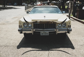 A vintage car with a large set of longhorns mounted on the front. The vehicle is parked on a paved area with buildings and trees in the background. Texas license plate is visible.