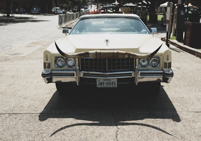 A vintage car with a large set of longhorns mounted on the front. The vehicle is parked on a paved area with buildings and trees in the background. Texas license plate is visible.