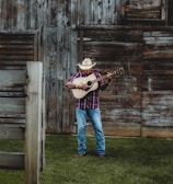 A man wearing a cowboy hat and a plaid shirt is playing an acoustic guitar in front of a rustic wooden barn. The ground is covered with lush green grass, and the barn features weathered wooden planks with a slightly open window.