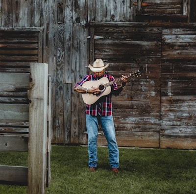 A man wearing a cowboy hat and a plaid shirt is playing an acoustic guitar in front of a rustic wooden barn. The ground is covered with lush green grass, and the barn features weathered wooden planks with a slightly open window.