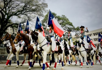 A group of people, dressed in coordinated outfits with cowboy hats, ride on horseback in a parade. They are holding flags with blue and white stars and one distinctive flag with a red, white, and blue pattern, resembling the Texas state flag. The horses are decorated with matching ribbons. The background includes large trees and a few buildings.