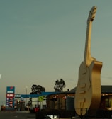 A large, golden guitar sculpture is prominently displayed in front of a building, with a recognizable fast food sign and a gas station in the background. Cars are parked nearby under a clear sky.