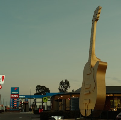 A large, golden guitar sculpture is prominently displayed in front of a building, with a recognizable fast food sign and a gas station in the background. Cars are parked nearby under a clear sky.