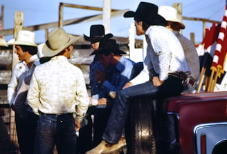 A group of men wearing cowboy hats and western-style clothing are gathered together outdoors. Some of them appear to be engaged in conversation, with one man sitting on the side of a vehicle. There are red, white, and black flags visible in the background, along with wooden fencing and a clear blue sky.