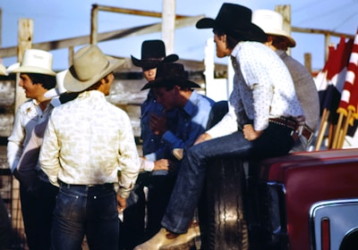 A group of men wearing cowboy hats and western-style clothing are gathered together outdoors. Some of them appear to be engaged in conversation, with one man sitting on the side of a vehicle. There are red, white, and black flags visible in the background, along with wooden fencing and a clear blue sky.