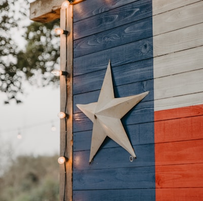 A wooden wall painted with the Texas flag design featuring vertical stripes of blue, white, and red, with a prominent white star in the middle of the blue section. The edge of the structure is adorned with small circular string lights. Trees and foliage are faintly visible in the blurred background, contributing to an outdoor setting.
