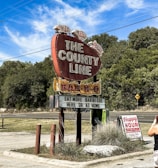 A large vintage-style red sign shaped like a brisket advertises a barbecue restaurant called 'The County Line.' The sign features pigs on top and a message encouraging patrons to 'eat more barbeque here or at home.' A smaller sign nearby announces 'Happy Hour' details. Surrounding the signs are patches of grass and trees under a clear blue sky with wispy clouds. A person with a camera is partially visible in the foreground.