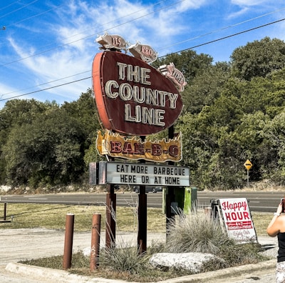 A large vintage-style red sign shaped like a brisket advertises a barbecue restaurant called 'The County Line.' The sign features pigs on top and a message encouraging patrons to 'eat more barbeque here or at home.' A smaller sign nearby announces 'Happy Hour' details. Surrounding the signs are patches of grass and trees under a clear blue sky with wispy clouds. A person with a camera is partially visible in the foreground.